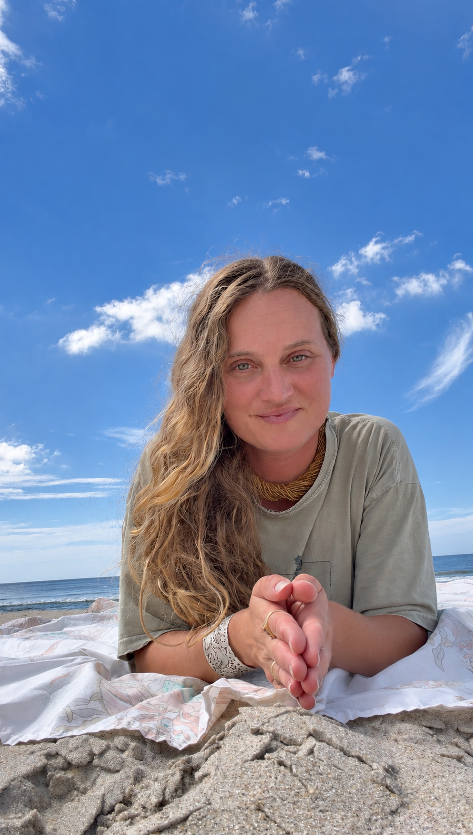Kir Grace resting on her elbows on a blanket in the sand, with the ocean behind her